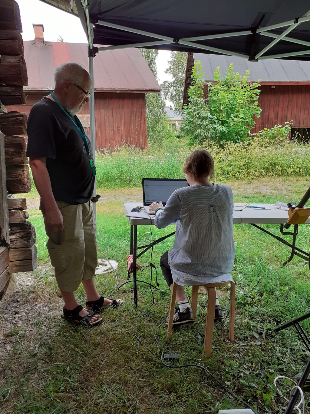 Kuvan vasemmassa laidassa mies, kuvan keskellä istuu nainen pöydän ääressä. Molemmat henkilöt ovat pop up-teltan alla, nainen tietokoneella. taustalla näkyy punamullattuja museoraitin rakennuksia, korkeaa heinää ja lehtipuita. Kuva on otettu kesällä pilvisenä päivänä.