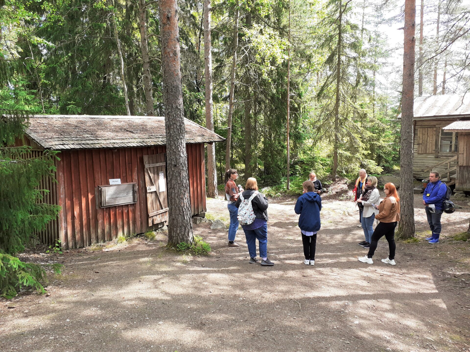 Kuvassa vasemmalla punainen lautarakenteinen savusauna, jonka vieressä seisoo ryhmä opastettavia, taka-alalla harmaa hirsirakennus.