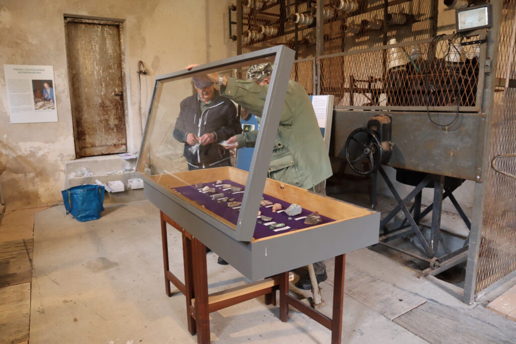 Kuva vanhan muuntamorakennuksen sisätiloista, jossa kaksi mieshenkilöä asettaa vitriiniin esille korukiviä.